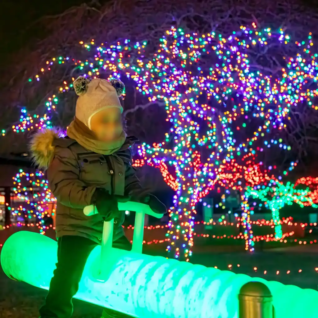 A child bundled in winter clothes happily riding a bright green LED seesaw, a perfect example of fun games with lights at an evening festival.