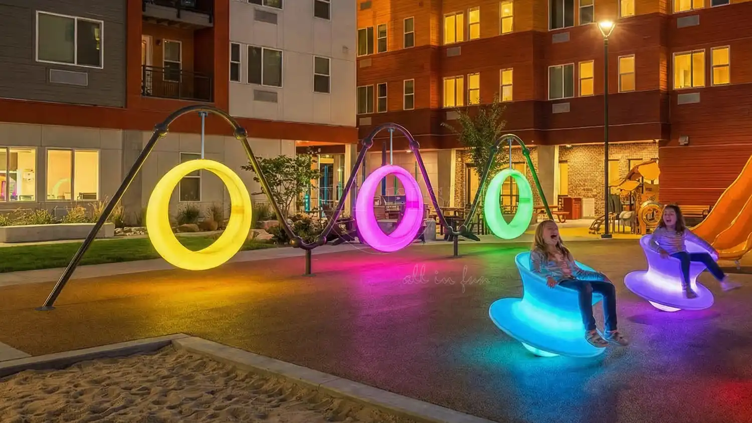 A dynamic nighttime view of a community playground featuring internally lit LED circle swings and glowing spinner chairs amidst modern residential buildings.