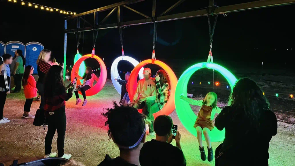 A night scene showing five colorful, glowing LED light up swings being used as outdoor entertainment games. Tourists take photos of children and adults on the swings in a festive setting