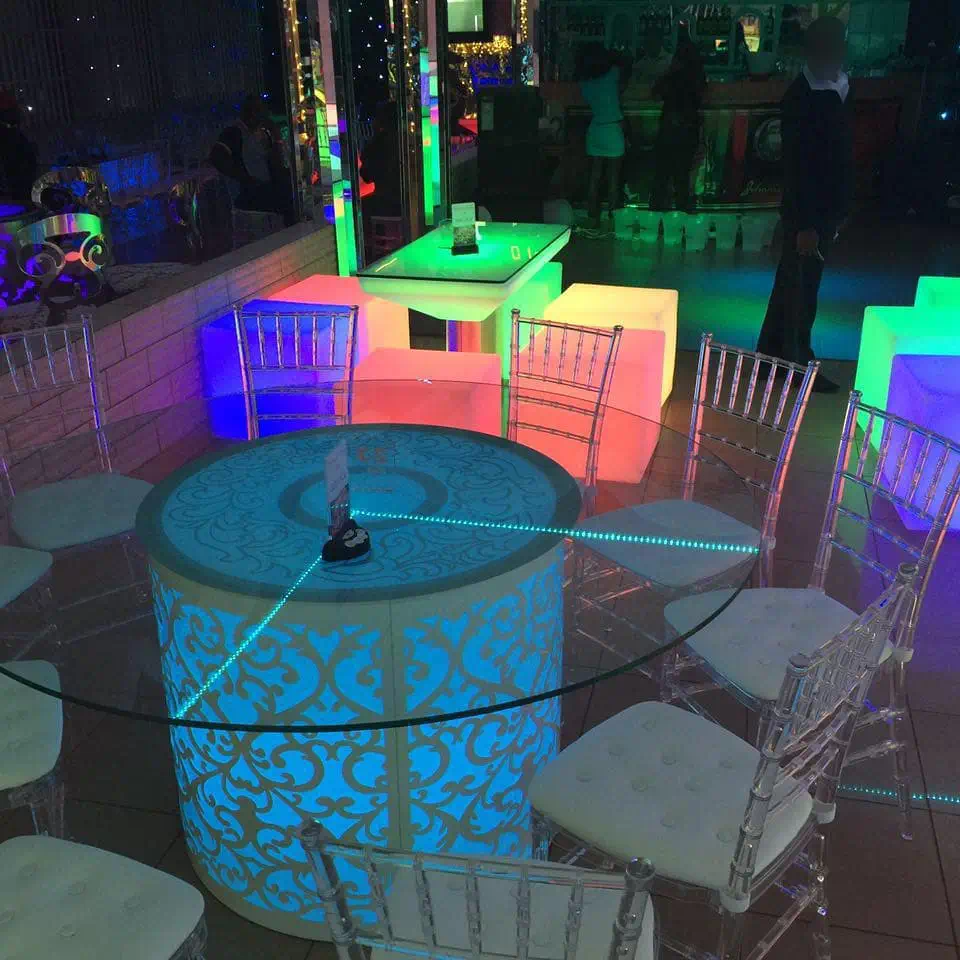 A vibrant event space with luminous furniture, featuring a round LED table and colorful Glow Cube Seats, surrounded by clear chairs