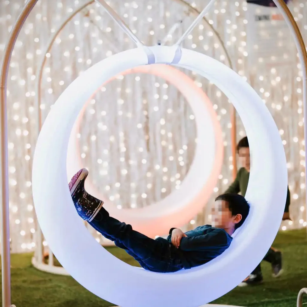Child relaxing comfortably inside a large, glowing white circular swing, a premium piece of LED Glow Furniture for events.