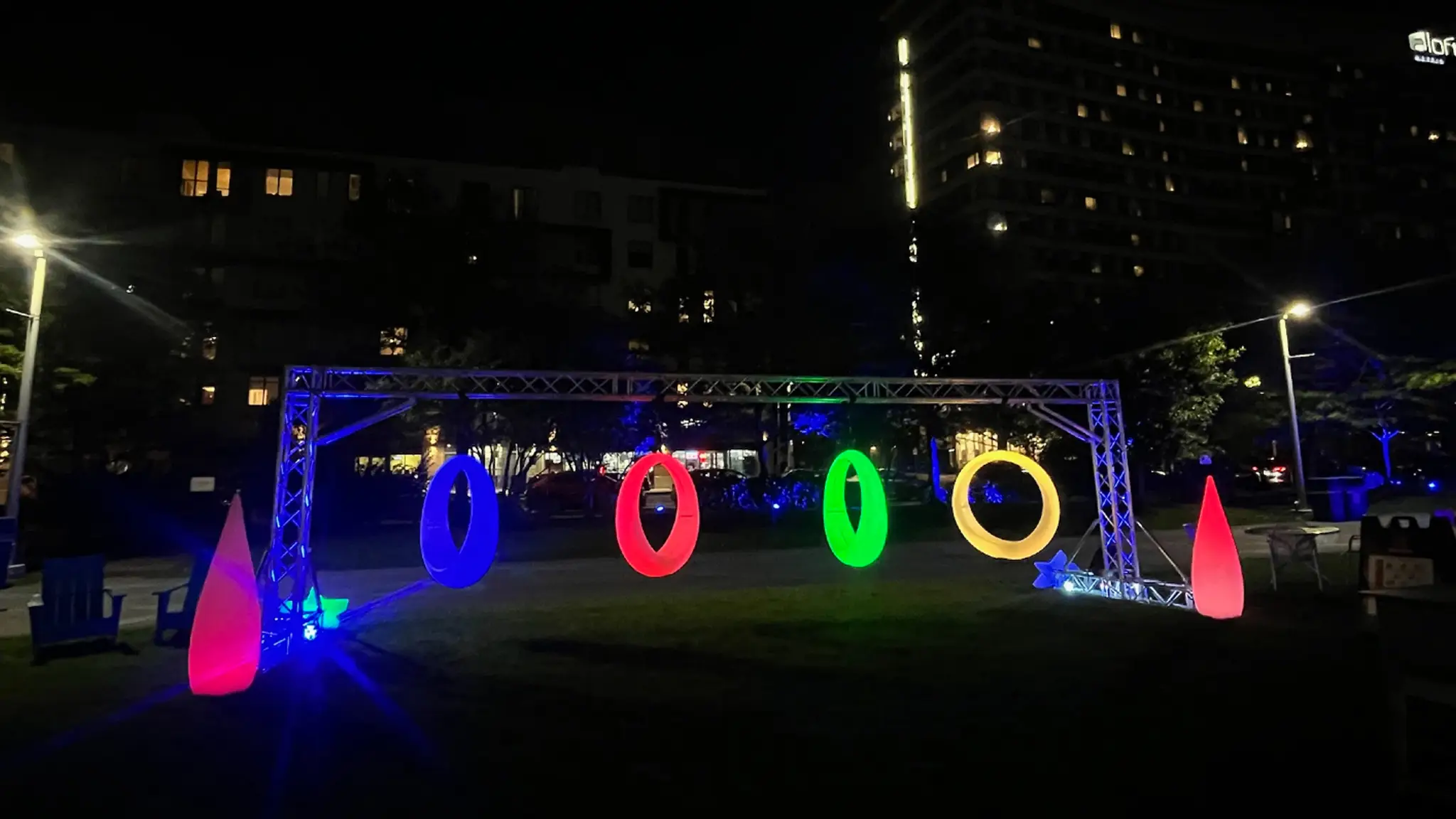 Four circular LED glow games swings in blue, red, green, and yellow, hanging from a metal truss at a night event.