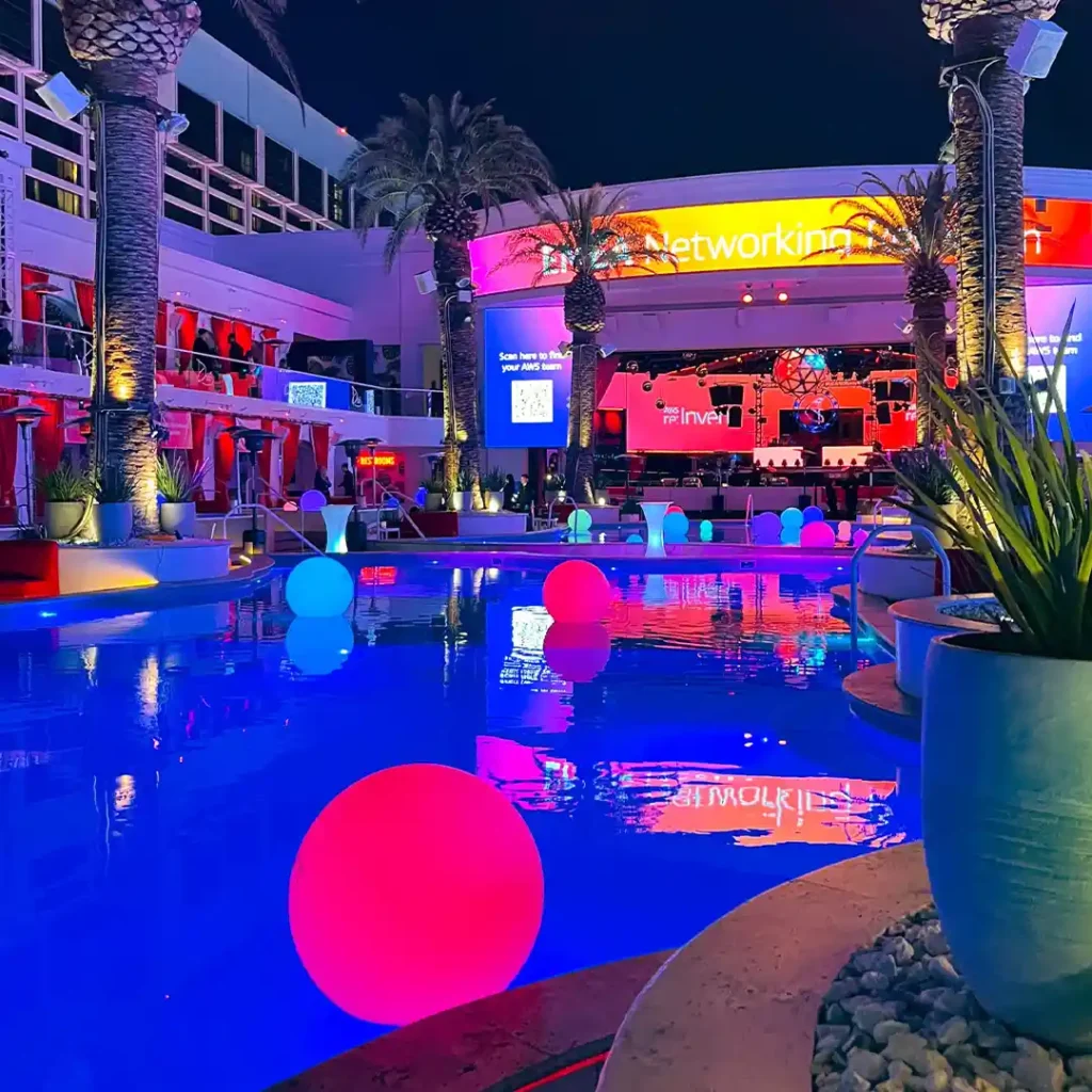 Glowing waterproof LED balls floating on a blue swimming pool at a nighttime party with neon palm trees.