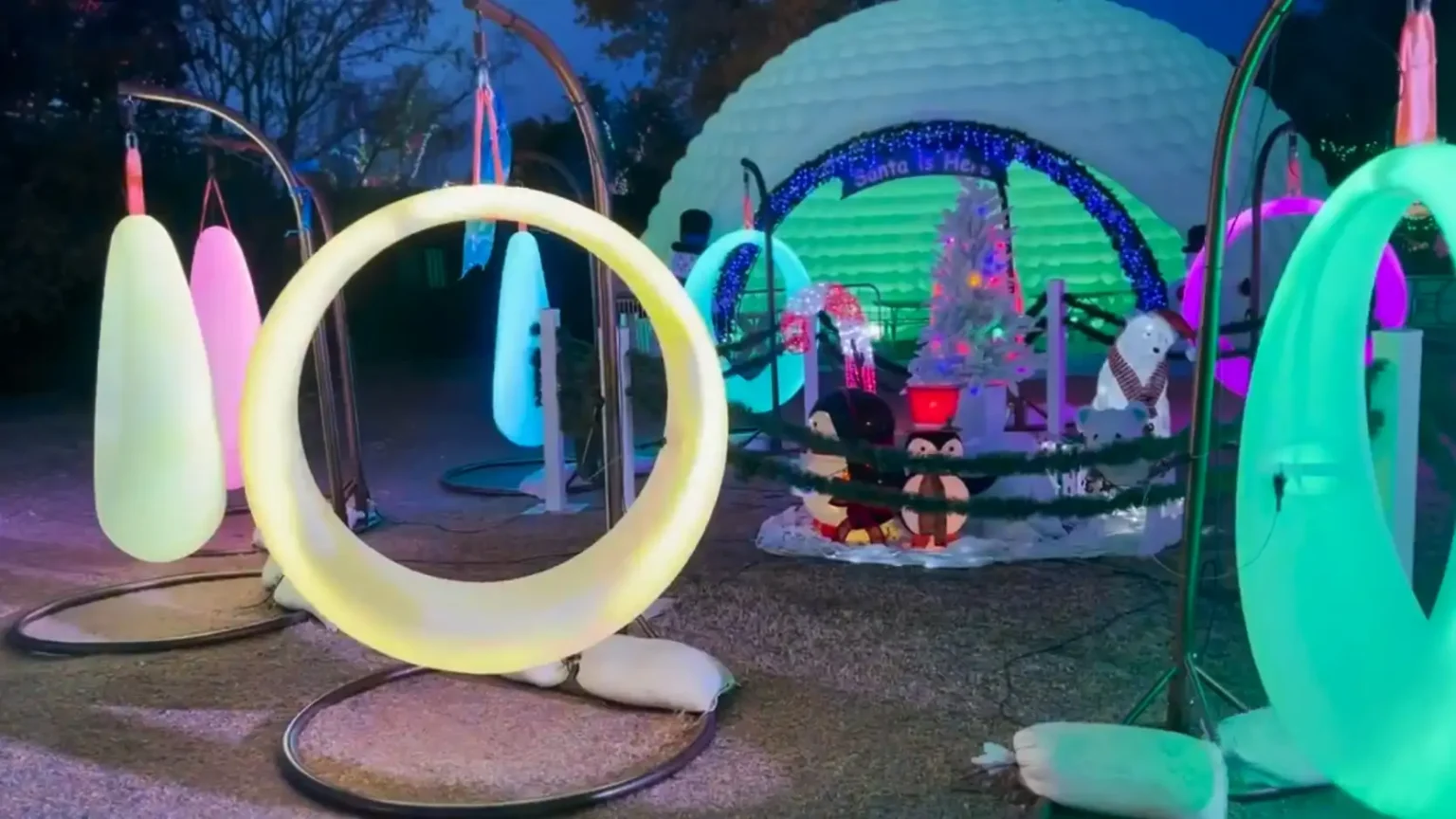 Several glowing LED circular swings hanging from frames, including white, yellow, and green lights, at an outdoor evening festival.