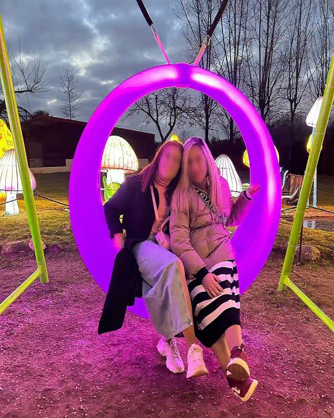 Two smiling girls enjoying a purple glowing LED outdoor furniture swing at a night park with lit mushroom decorations
