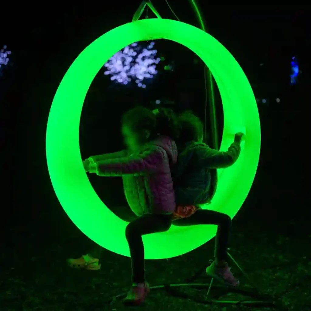 Two-young-girls-sitting-back-to-back-inside-a-large,-illuminated-green-Glow-Circular-Swing-in-a-dark-outdoor-setting