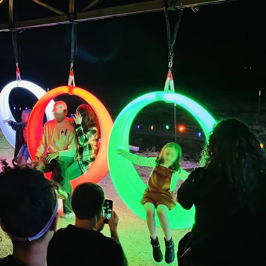 night-scene-showing-five-colorful-glowing-LED-light-up-swings-being-used-as-outdoor-entertainment-games.-Tourists-take-photos-of-children-and-adults-on-the-swings-in-a-festive-setting