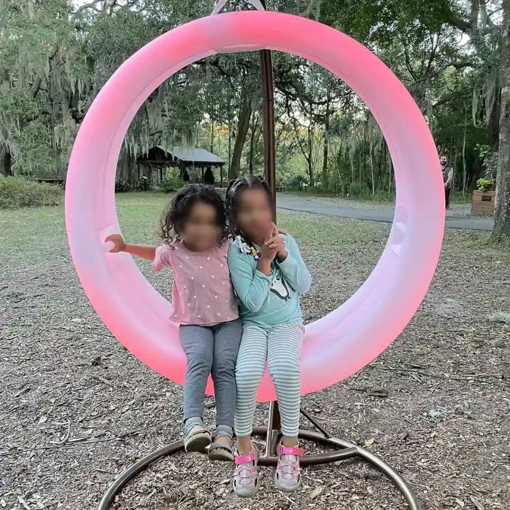 two-young-children-sitting-and-smiling-together-inside-a-large,-glowing-pink-Glow-Circular-Swing-on-a-stand-outdoors