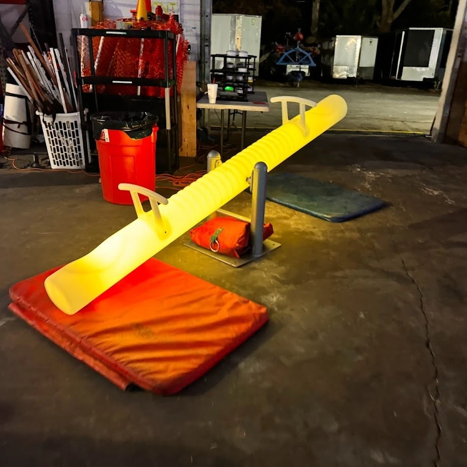 A brightly illuminated yellow LED seesaw, a piece of furniture glow event rental equipment in Florida, is positioned in a cluttered warehouse setting in front of various objects