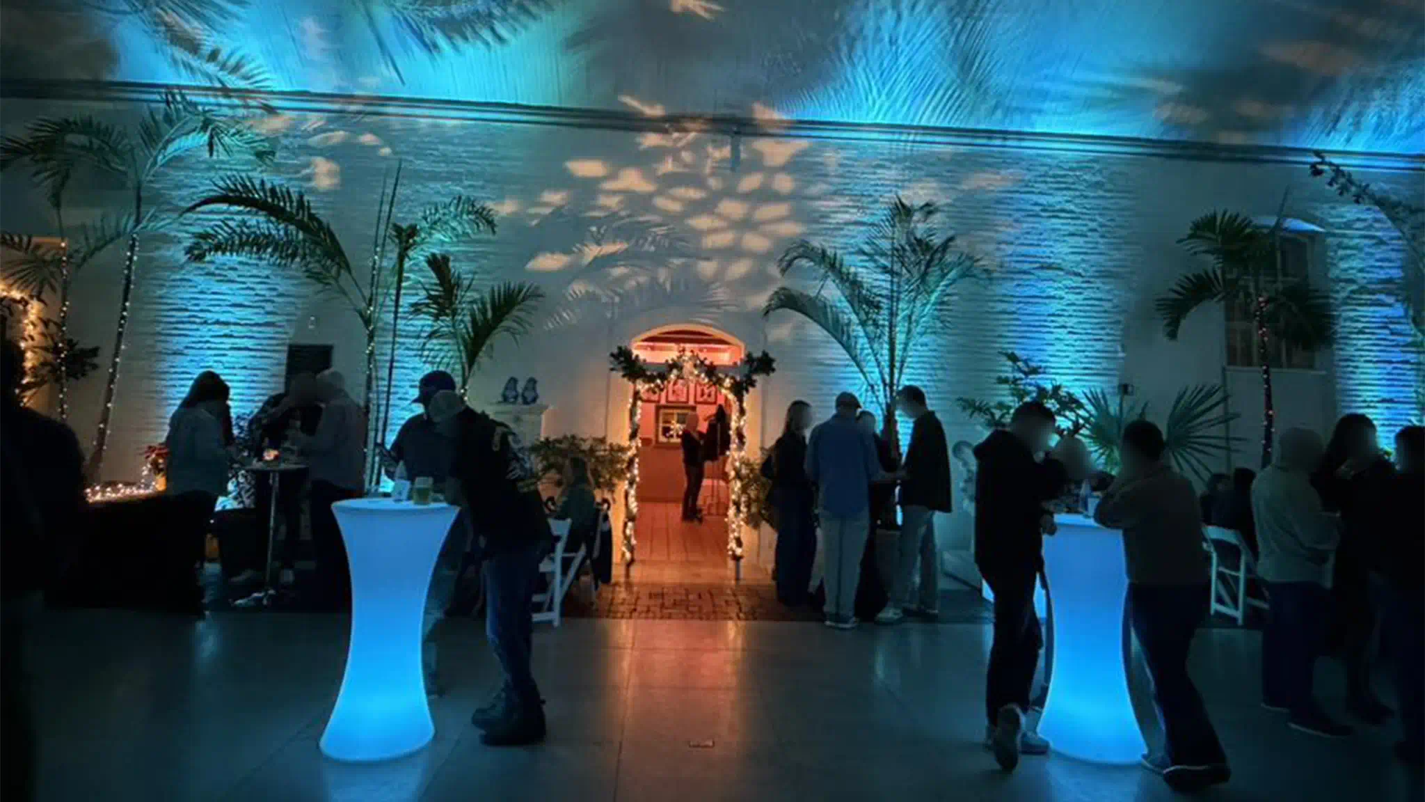 A group of people standing around hour-glass shaped LED lighted tables that are internally lit with a vibrant blue glow at a cocktail party