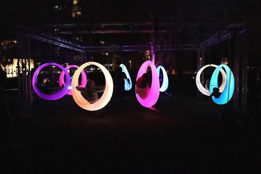 Colorful interactive furniture led lighting display in Hengelo Netherlands, featuring eight multi-color LED swings with adults and children playing at night
