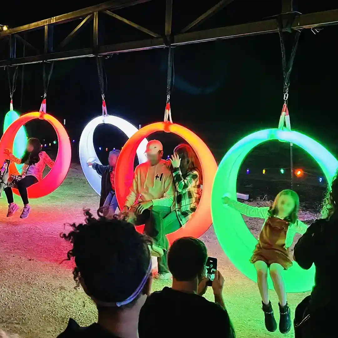 Des balançoires lumineuses à LED sont utilisées comme jeux de divertissement en plein air. Des touristes prennent des photos d'enfants et d'adultes sur les balançoires dans une ambiance festive.